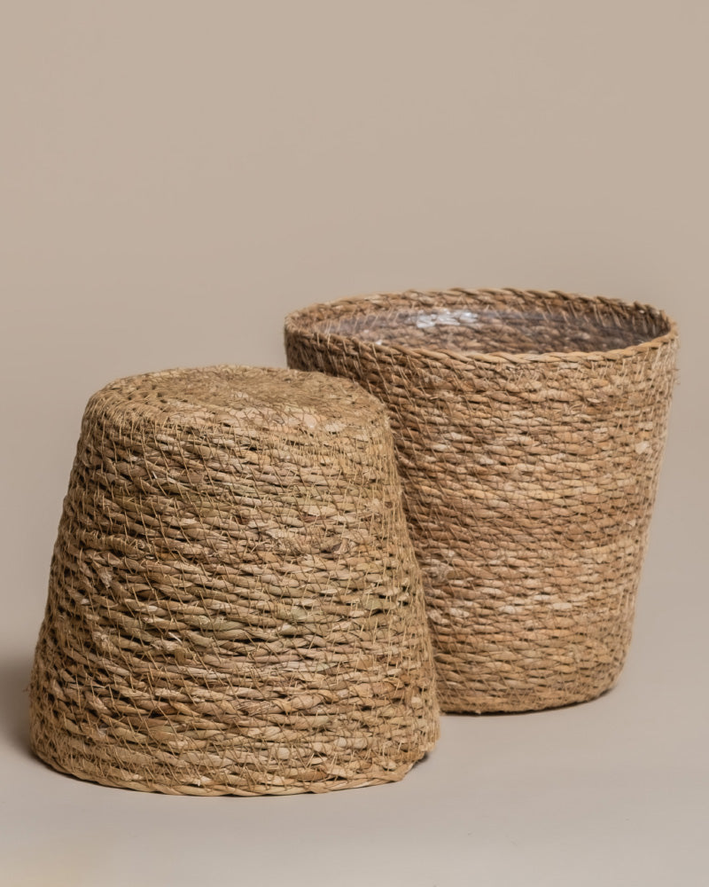 Der Pflanzkorb aus Seegras (Kesat | 16 cm ⌀) zeigt in zwei handgeflochtenen Varianten, aufrecht stehend oder seitlich liegend, seine dekorative, natürliche Struktur vor schlicht beigem Hintergrund.