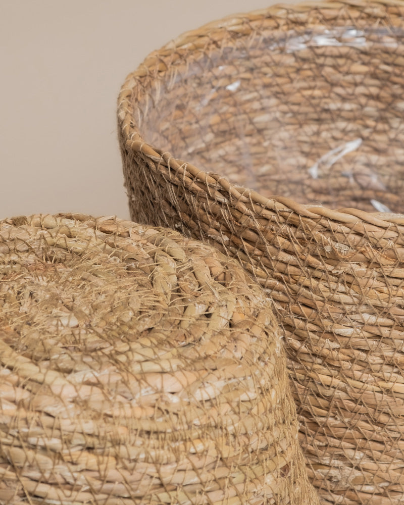 Nahaufnahme von zwei Pflanzkörben aus Seegras (Kesat | 16 cm ⌀) - einer aufrecht, einer auf der Seite - in naturbraun mit durchsichtigem Kunststofffutter, wodurch sie wasserdicht und ideal für Zimmerpflanzen sind.