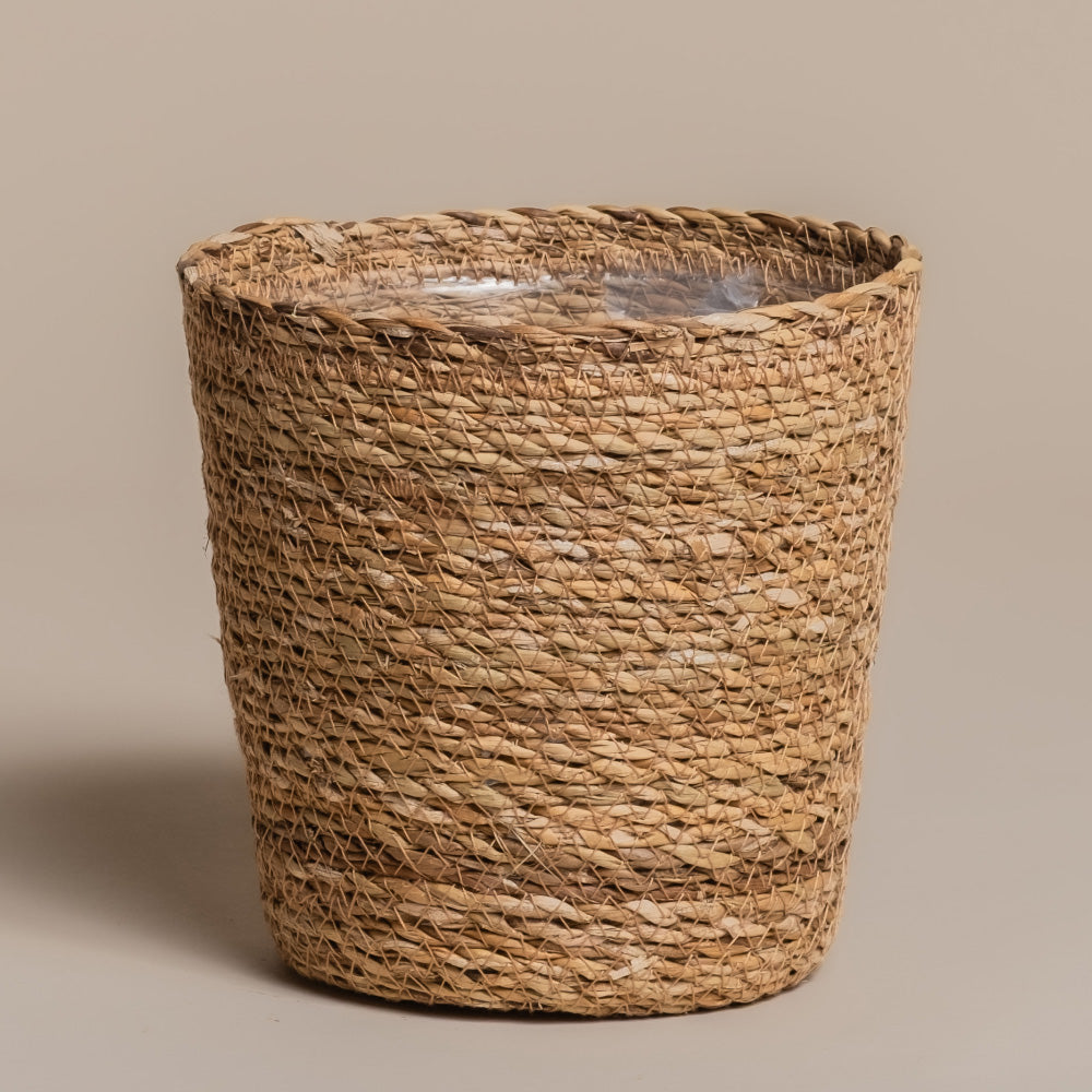 Der Pflanzkorb aus Seegras (Kesat | 18 cm ⌀) ist handgemacht, zylindrisch, mit leicht unregelmäßigem Rand und verfügt über ein wasserdichtes, transparentes Kunststofffutter. Präsentiert vor schlicht beigem Hintergrund.