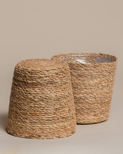 Zwei handgefertigte "Pflanzkorb aus Seegras (Kesat | 18 cm ⌀)": Einer steht aufrecht mit wasserdichter Kunststoffauskleidung, der andere liegt umgedreht und zeigt den flachen Boden - beide vor schlichtem beigem Hintergrund.