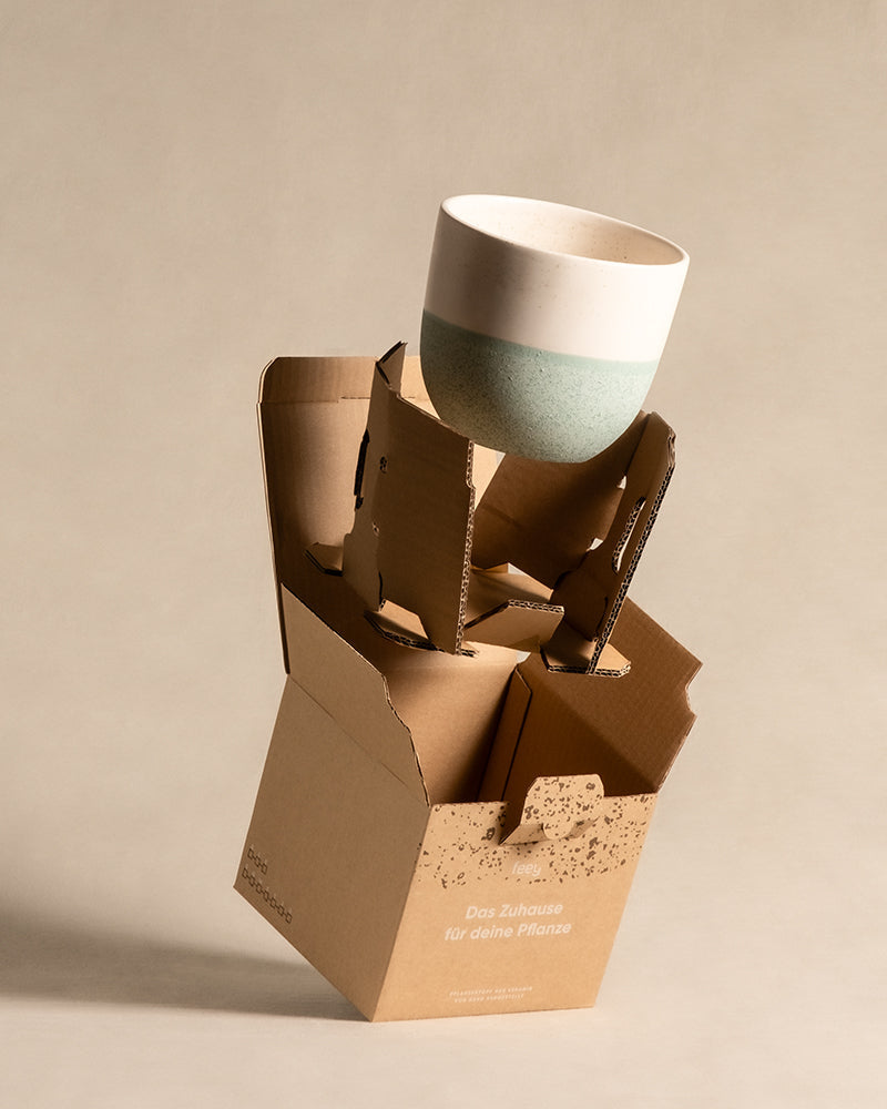 Das Keramik-Topfset „Variado“ (16, 14, 7) mit handgefertigtem Design in Weiß und Hellgrün wird über einem offenen, umweltfreundlichen Karton präsentiert. Die Verpackung mit deutschem Etikett enthält praktische Stützen im Inneren, die die Töpfe sicher halten.