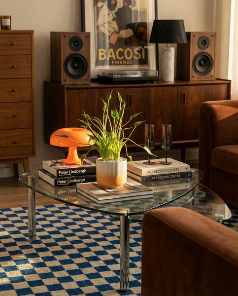 Ein gläserner Couchtisch mit einer Alocasia 'Tiny Dancer' von feey, Büchern und einer orangefarbenen Lampe steht auf einem blau karierten Teppich in einem gemütlichen Wohnzimmer mit Holzmöbeln, Lautsprechern, gerahmter Kunst und warmem Sonnenlicht, das hereinströmt.