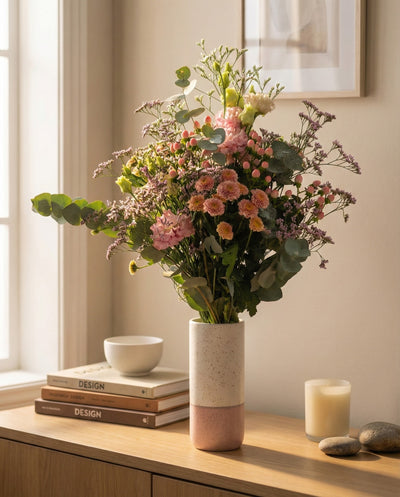 Der Morgenlichtstrauß aus rosa und lila Herbstblumen mit Grünzeug in einer weiß-rosa Vase steht auf einem Holztisch neben Büchern, einer Kerze und einer Schale, während sanftes Sonnenlicht den Raum durch das Fenster erhellt.