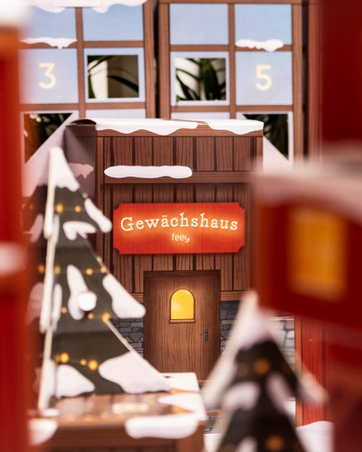 Ein gemütliches, verschneites Holzhaus mit dem leuchtenden Schild "Gewächshaus feey" steht zwischen Papierbäumen und verschneiten Dächern und weist mit seinen nummerierten Fenstern auf den Pflanzen-Adventskalender im Hintergrund hin.