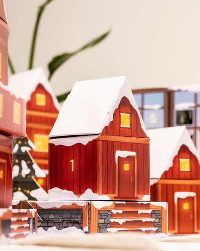 Feiern Sie die Saison mit dem Pflanzen-Adventskalender - ein festliches rotes Holzhaus mit der Aufschrift "1", leuchtenden Fenstern und einem verschneiten Dach, eingebettet zwischen Topfpflanzen, ähnlichen Häusern und Weihnachtsdekoration.
