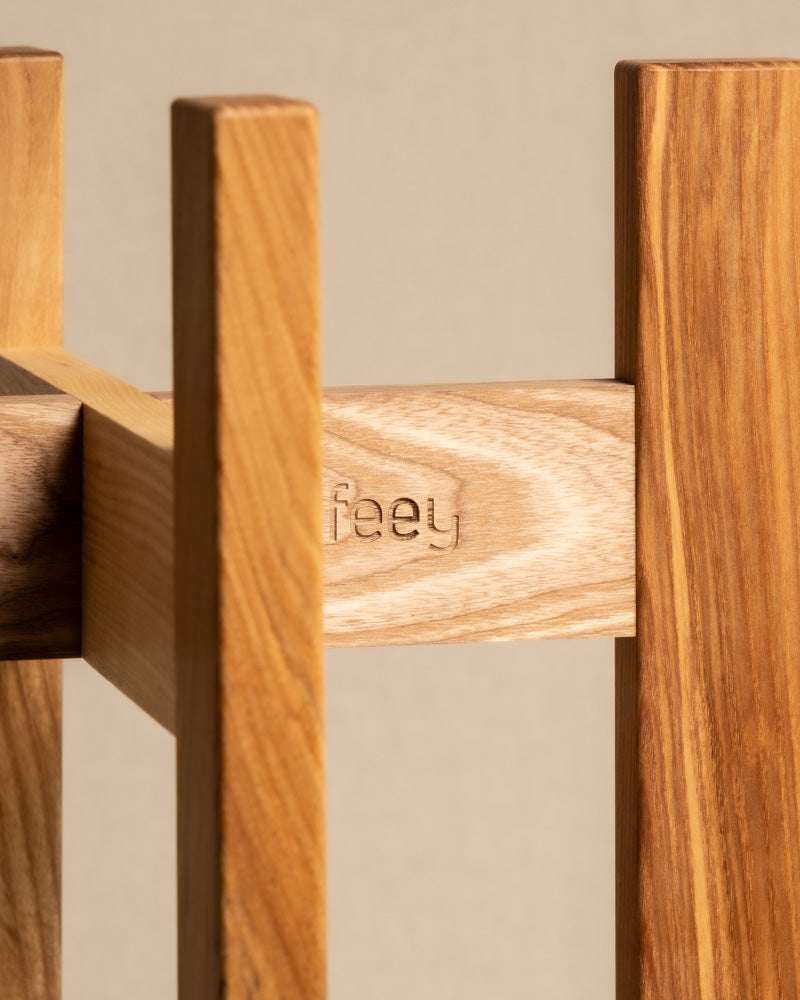 Nahaufnahme einer Holzkonstruktion aus Eschenholz mit hellbrauner Oberfläche, auf einer der vertikalen Lamellen ist das Wort „feey“ eingeprägt. Der sanft verschwommene Hintergrund betont die Textur und Maserung und erinnert an den Pflanzenständer (Stelion | 16 cm Ø).