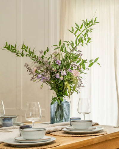 Das Esstischset Purpurpoesie mit hellblauen Tellern, Schalen, Weingläsern und gewebten Platzsets strahlt Eleganz aus. Eine hohe Vase mit üppigen grünen Blättern und violetten Blumen bildet das Herzstück, während sanftes natürliches Licht den Raum füllt.