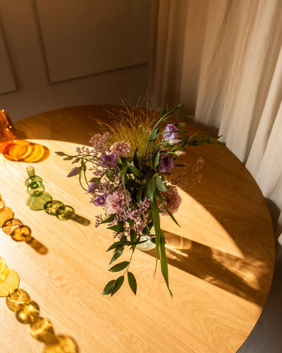 Der Purpurpoesie-Strauß in Lila und Rosa mit grünen Blättern ruht in einer Glasvase auf einem runden Holztisch neben buntem Glasdekor, das in Sonnenlicht getaucht ist - ein Fest der natürlichen Anmut.
