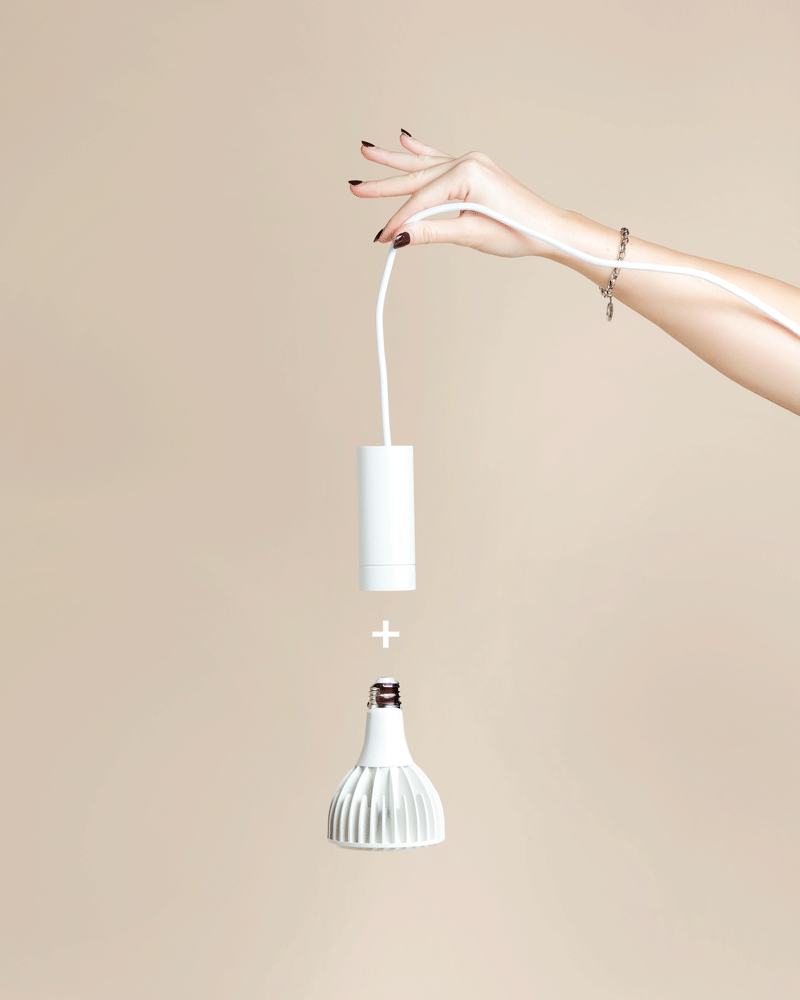 Eine Hand mit lackierten Nägeln hält eine weiße Pendelleuchte 'Corda' über einer weißen Glühbirne, mit einem Pluszeichen dazwischen, vor einem beigen Hintergrund.