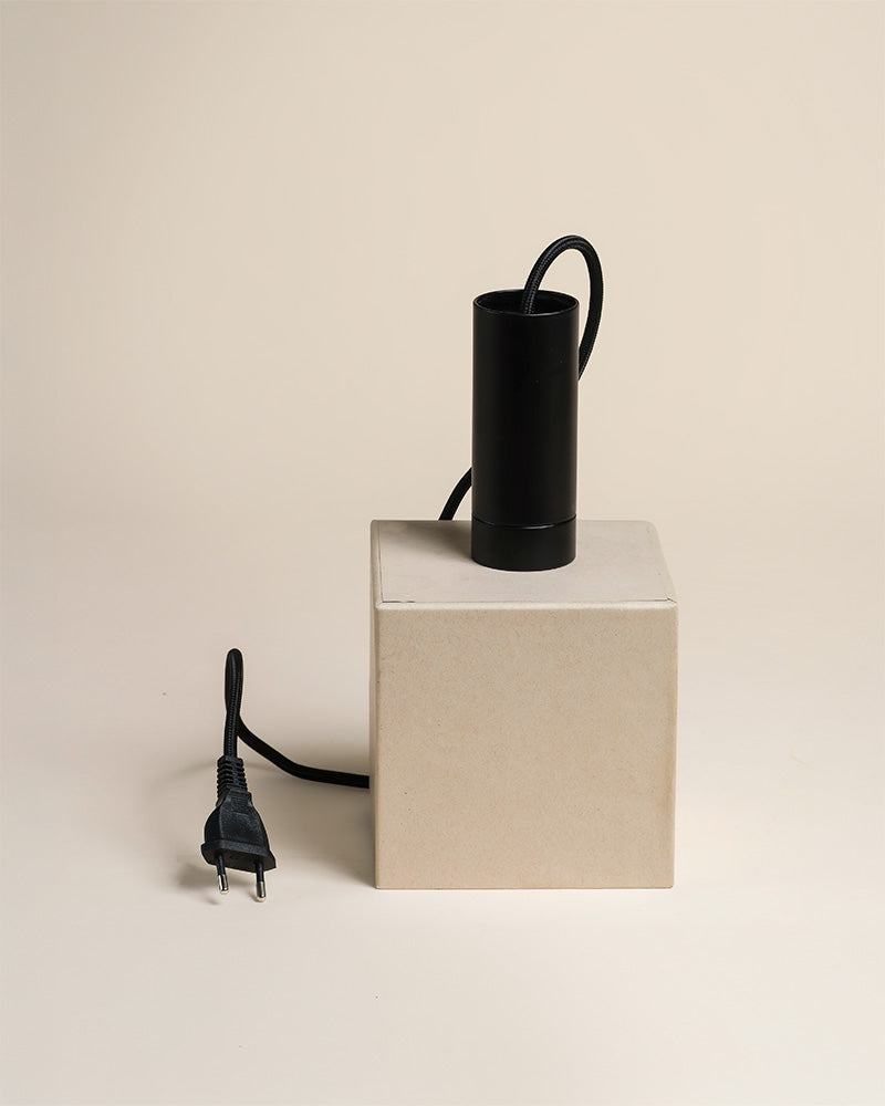 Eine schwarze zylindrische Pendelleuchte "Corda" mit Netzkabel und Stecker ruht auf einem beigen rechteckigen Kasten vor einem schlichten hellen Hintergrund.