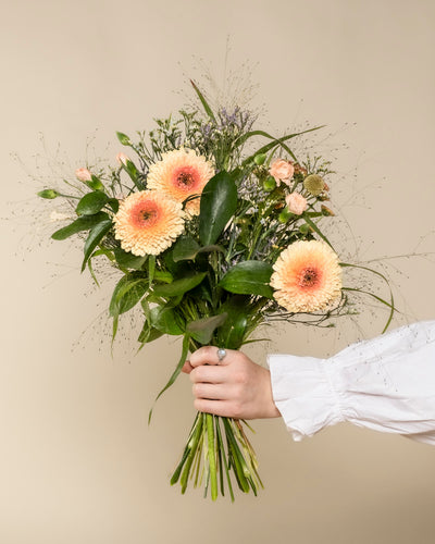 Eine Hand in einem weißen Ärmel hält den Überraschungs-Blumenstrauß mit hellpfirsichfarbenen Gerbera, kleinen pfirsichfarbenen Rosen, grünen Blättern und zarten Füllern - ein charmantes Geschenk vor einem beigen Hintergrund.