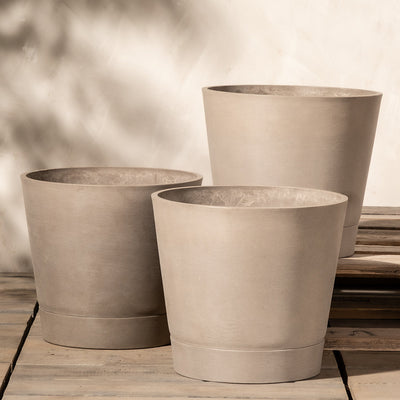 Drei große, beige Pflanzkübel-Set Venice (3x 30) in unterschiedlichen Höhen sind in einer Gruppe auf einer Holzfläche angeordnet. Den Hintergrund bildet eine helle Wand mit weichen, gesprenkelten Blätterschatten, die der Szene eine ruhige, natürliche Atmosphäre verleihen. Jeder Pflanzkübel verfügt über ein effizientes Drainage-System für optimale Pflanzengesundheit.