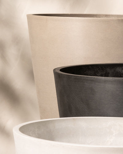 Drei große, strukturierte Pflanzkübel – gefertigt aus Recycling-Kunststoff – werden hintereinander präsentiert. Der vordere Pflanzkübel ist weiß, der mittlere schwarz und der hintere beige. Der Hintergrund weist einen subtilen, verschwommenen Farbverlauf von hell nach dunkel auf, der die Formen und Strukturen des Pflanzkübel-Sets Venedig (3x 30) hervorhebt.