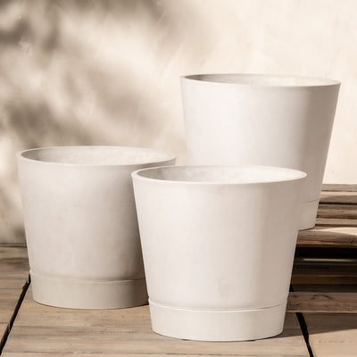 Drei große, weiße Pflanzkübel-Sets Venice (3x 30) stehen versetzt auf einer Holzfläche. Die Töpfe haben ein minimalistisches Design mit glatten, leicht konischen Seiten, die subtile Schatten werfen. Der Hintergrund verfügt über ein innovatives Drainagesystem und besticht durch sanfte, neutrale Töne mit gesprenkeltem Licht.
