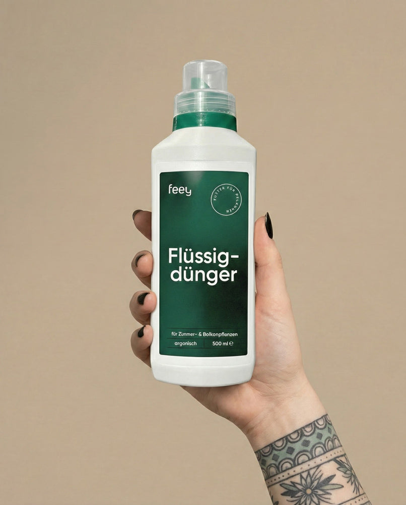 Eine Hand mit schwarzem Nagellack und einer floralen Tätowierung hält eine weiße Flasche Flüssigdünger vor einem beigen Hintergrund. Die Flasche für Zimmerpflanzen hat ein grünes Etikett und einen passenden grünen Verschluss.