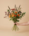 Hand hält den Blumenstrauß Sonnenwende in der Größe Classic mit oranger Färberdistel, rostroten Chrysanthemen und dunklen Beeren vor sandfarbenem Hintergrund