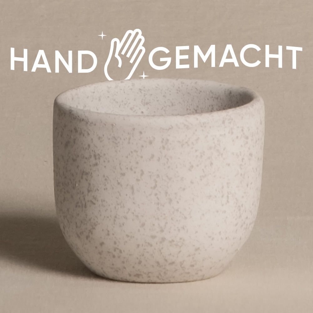 Ein Bild einer kleinen, strukturierten, weißen Keramiktasse vor einem hellbeigen Hintergrund. Über der Tasse steht das Wort „HANDGEMACHT“ in Großbuchstaben, mit einem Handsymbol zwischen „HAND“ und „GEMACHT“. Die natürliche Note verleiht dem Becher Charme und passt perfekt zu einem Keramik-Babypflanzen-Topf „Direito“.