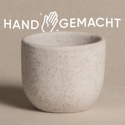 Ein Bild einer kleinen, strukturierten, weißen Keramiktasse vor einem hellbeigen Hintergrund. Über der Tasse steht das Wort „HANDGEMACHT“ in Großbuchstaben, mit einem Handsymbol zwischen „HAND“ und „GEMACHT“. Die natürliche Note verleiht dem Becher Charme und passt perfekt zu einem Keramik-Babypflanzen-Topf „Direito“.