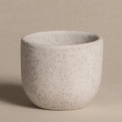 Eine kleine, runde, weiße Keramiktasse mit gesprenkelter Textur steht auf einem beigen Hintergrund. Die Tasse hat eine glatte, matte Oberfläche und strahlt ein minimalistisches Design mit einem natürlichen Touch aus. Perfekt als Keramik Babypflanzen-Topf 'Direito'.