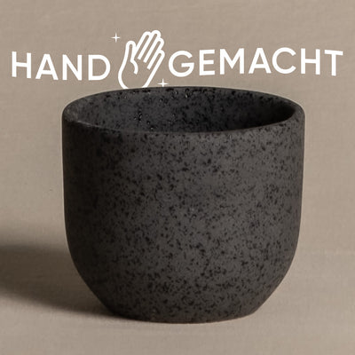 Ein dunkler, strukturierter Keramik-Babypflanzen-Topf „Direito“ wird auf einer glatten Oberfläche ausgestellt. Über dem Topf steht das Wort „HAND“ in weißen Großbuchstaben, mit einer Handillustration und Glitzer, gefolgt von „GEMACHT“, was bedeutet, dass der Artikel handgefertigt ist.