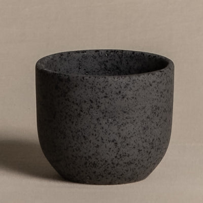 Ein kleiner, runder Keramik-Babypflanzen-Topf „Direito“ vor einem neutralen, beigen Hintergrund. Der schwarz gesprenkelte Keramikbecher hat ein glattes und leicht konisches Design ohne Henkel und zeigt einen minimalistischen Stil mit einer natürlichen Note.