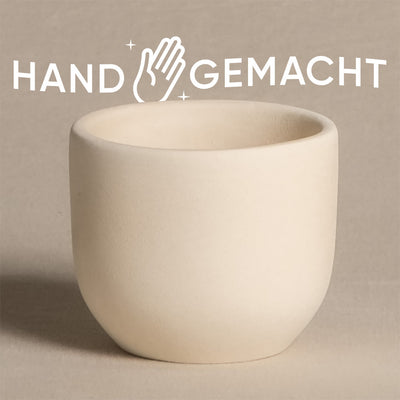 Eine einfache, weiße Keramiktasse steht vor einem neutralen Hintergrund. Darüber steht der Text „HAND GEMACHT“ mit einer stilisierten Handgrafik zwischen den Wörtern. Der Keramik-Babypflanzen-Topf „Direito“, der problemlos als Keramik-Übertopf für Babypflanzen dienen könnte, hat ein glattes, minimalistisches Design und eine unglasierte, matte Oberfläche.